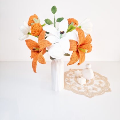bouquet de lys au crochet orange et blancs avec fleurs rondes crème et feuillage vert dans un vase blanc, sur napperon beige